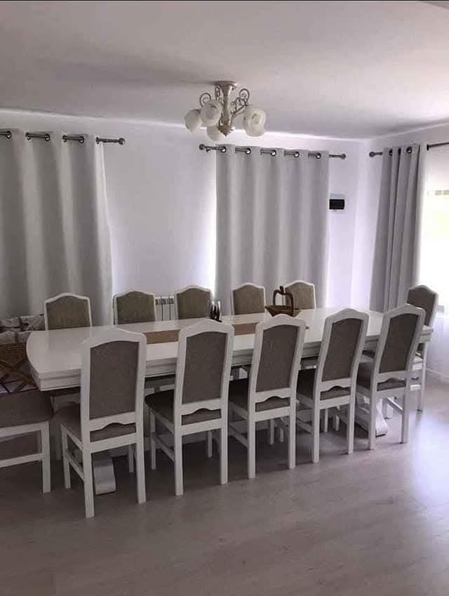 Masă lungă albă cu scaune albe tapițate în bej, într-o cameră cu perdele deschise la culoare